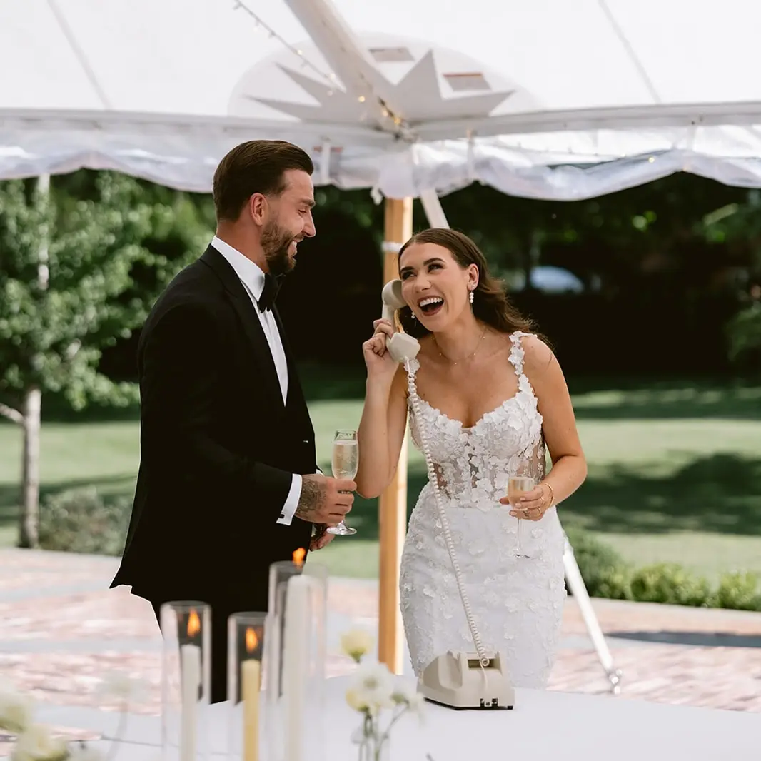 Laughing bride with the handset at her ear, next to the groom, under a tent in the garden