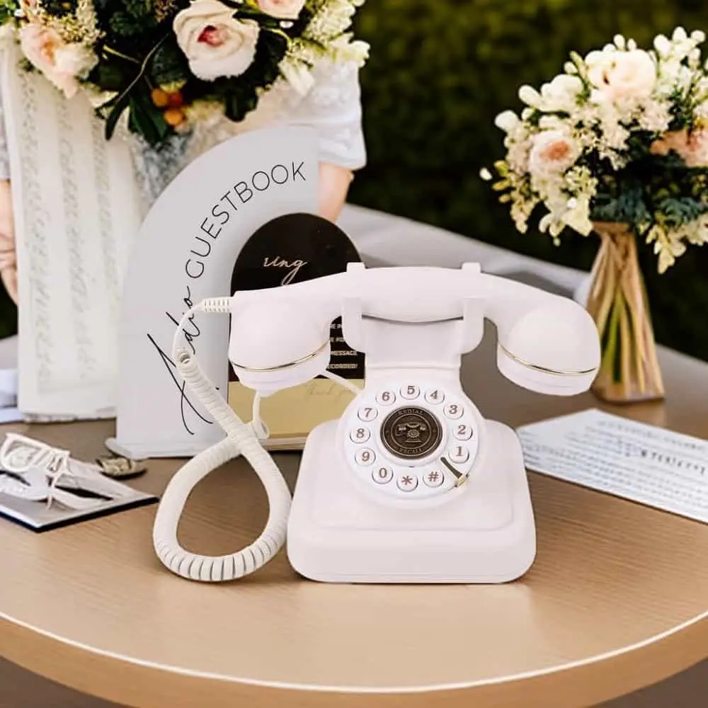 Белый ретро телефон Elegance70 White на круглом столе рядом с табличкой гостевой книги и цветами
