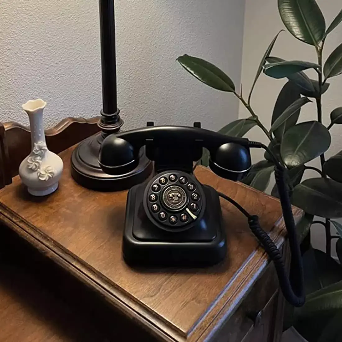 Black rotary phone on a wooden dresser, next to a lamp and a white vase