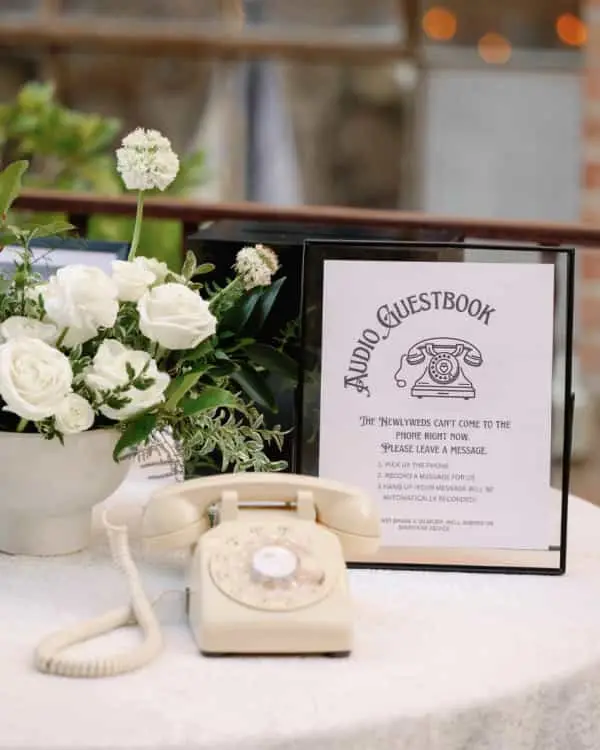 Cream rotary phone on a round table, next to an Audio Guestbook sign and white roses